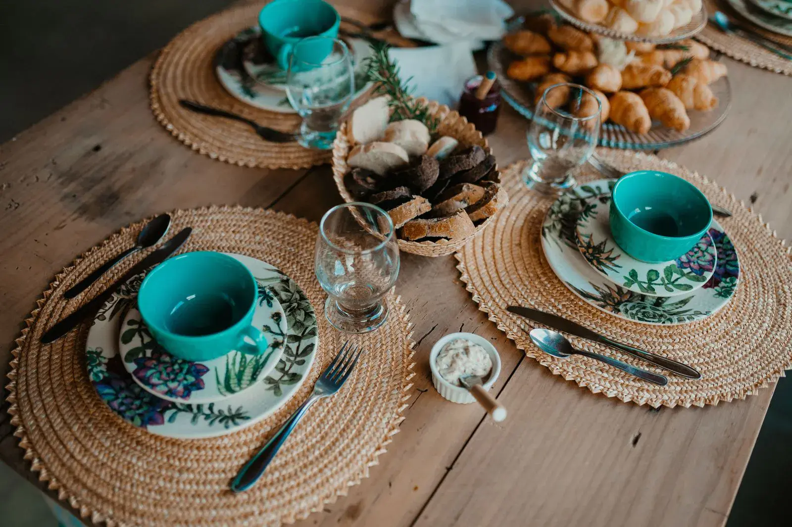 Table de petit-déjeuner dressée avec des tasses turquoise, des viennoiseries et des sets en osier