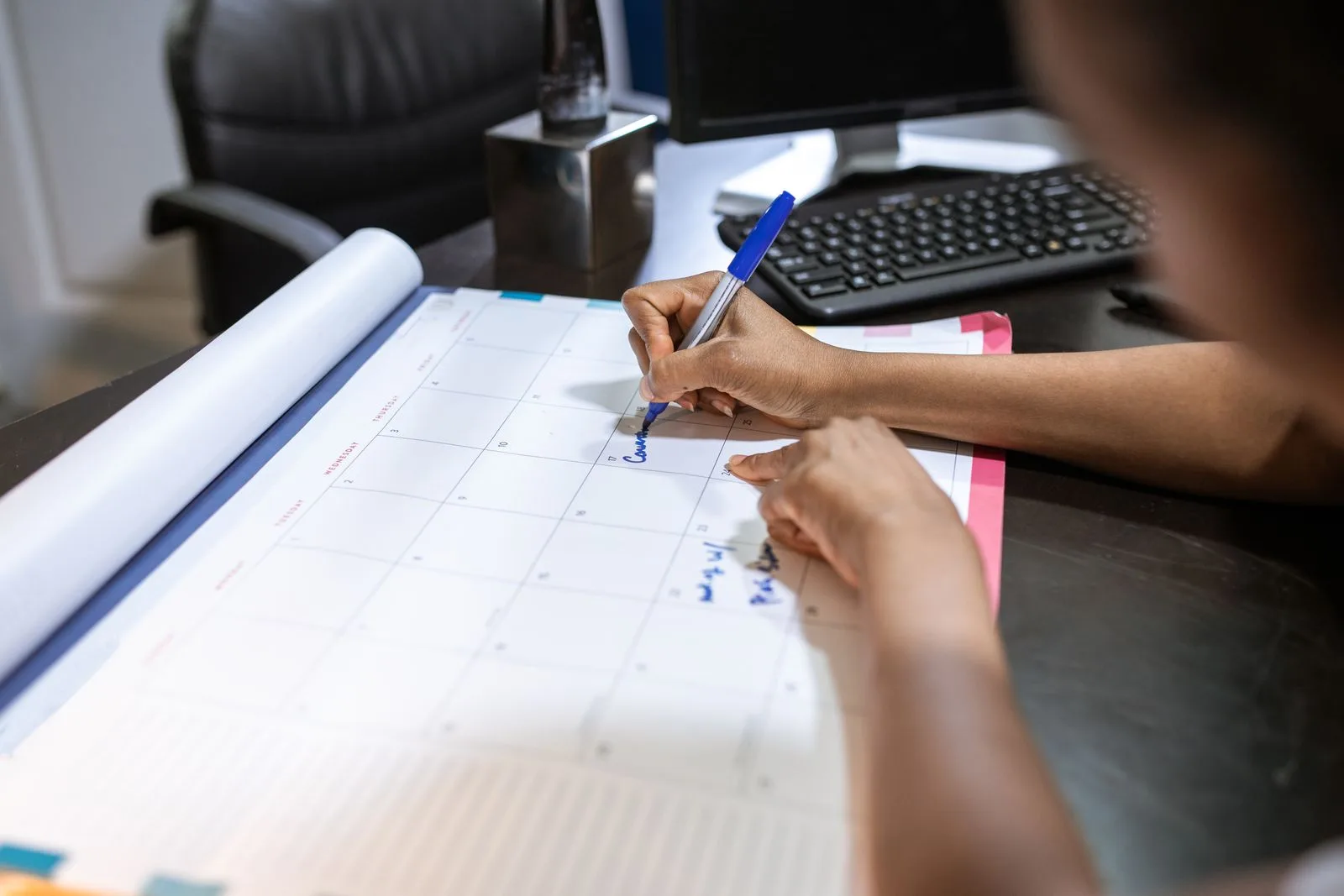 Main notant des réservations au stylo sur un grand calendrier mensuel posé sur un bureau